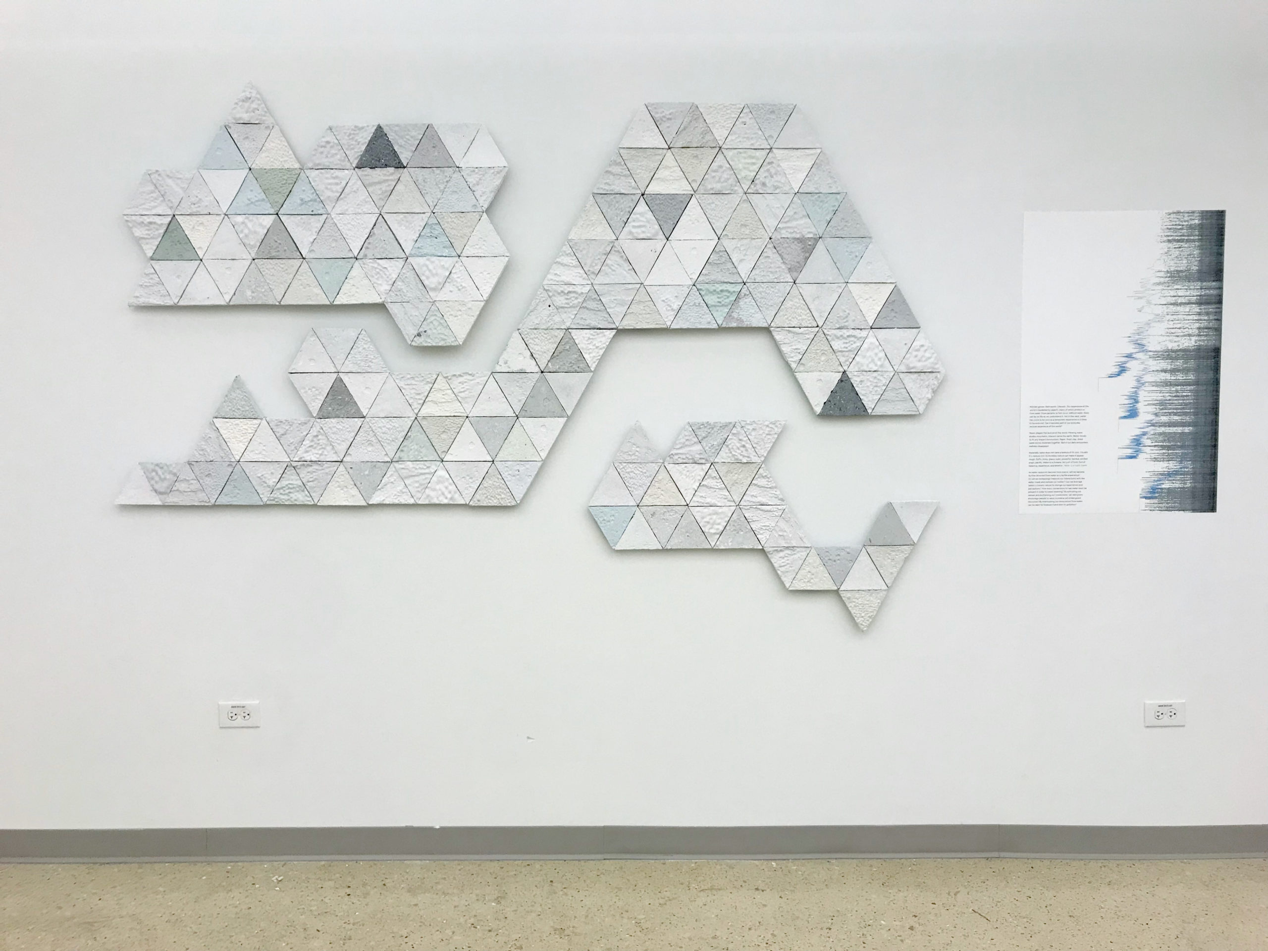 Three irregularly shaped but modular panels mounted to a wall with a poster to the right of them. The panels are made from paper and lightly colored white, green, blue, and gray