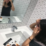 and Indian woman looking in the mirror of a bathroom mirror as she puts a sticker with a cut-out over her eye. A sheet of stickers is next to her on the counter