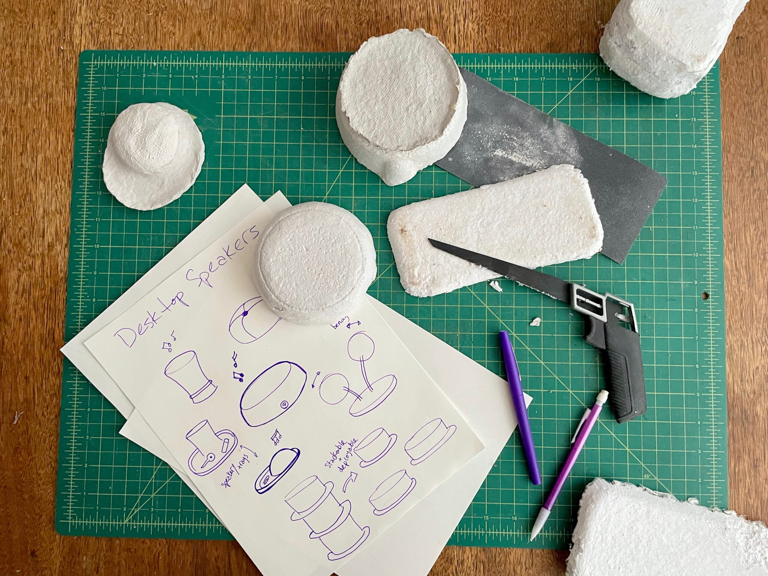 sketches and cai board and clay on top of a green cutting mat with tools such as pencils and saws