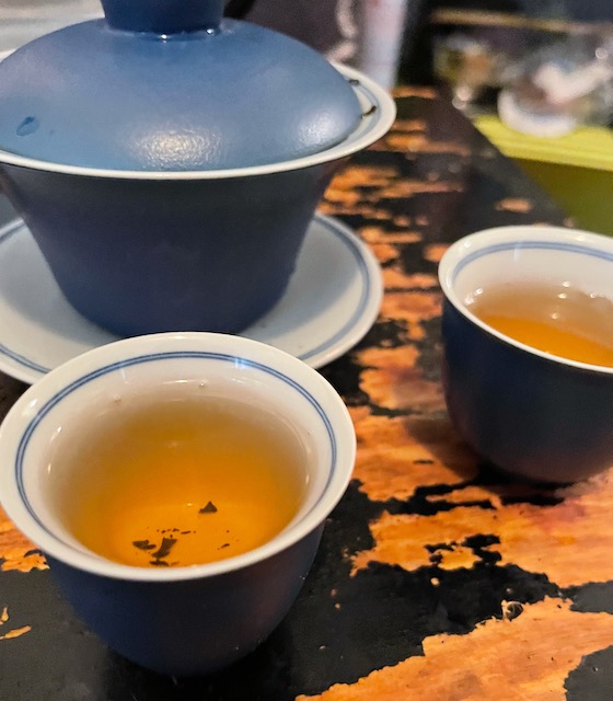 tea in two small, blue and white tea cuprs, poured from a gaiwan with the same colors, a gaiwan is a traditional chinese brewing vessel of a cup with a small lid on a saucer